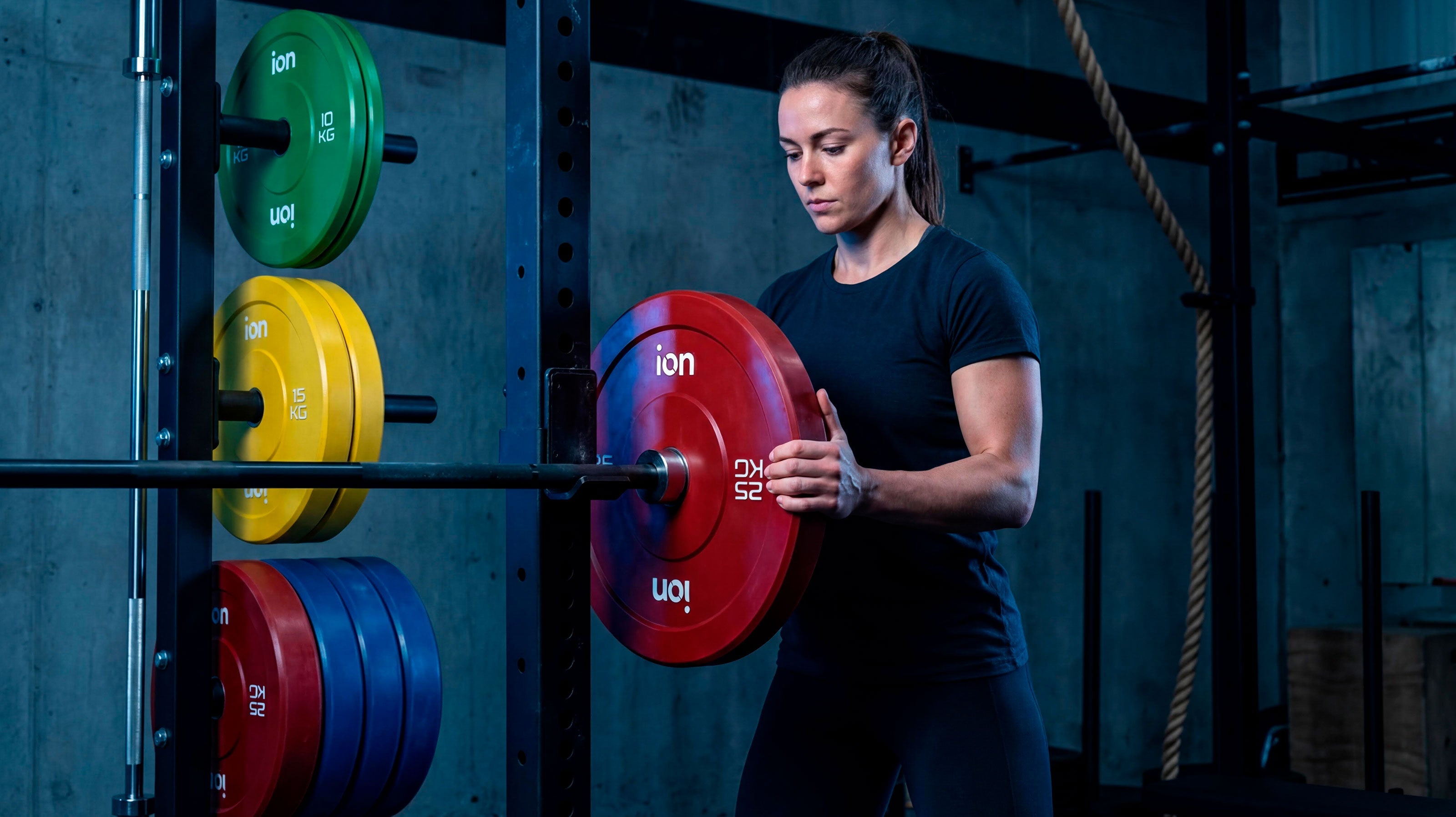 Una mujer desliza un disco de pesas rojo de la marca ION de 25 kg en una barra de pesas. La barra a su vez reposa sobre un rack. De fondo se ven más discos de peso de diferentes colores y pesos.