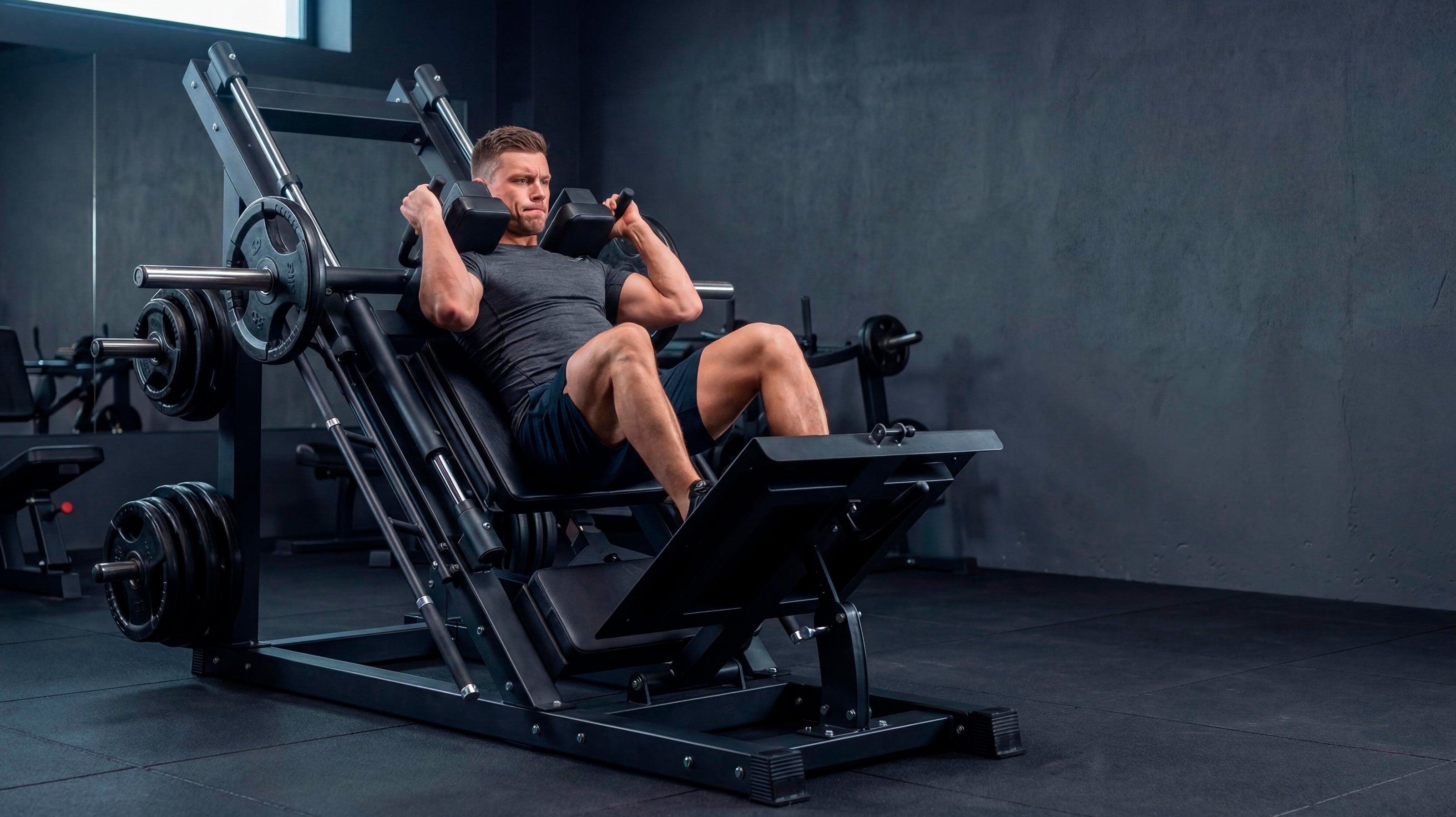 Un hombre pone cara de esfuerzo mientras realiza una sentadilla. Realiza el ejercicio en una máquina dual de prensa de piernas y sentadilla. De fondo se ven más máquinas de musculación.